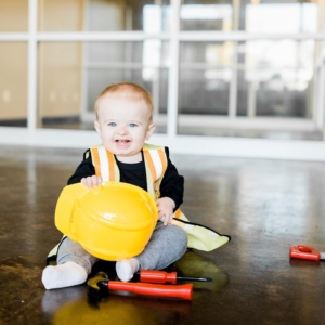 A baby wears a construction vest and grips a yellow hard hat with tools sitting in front of him.